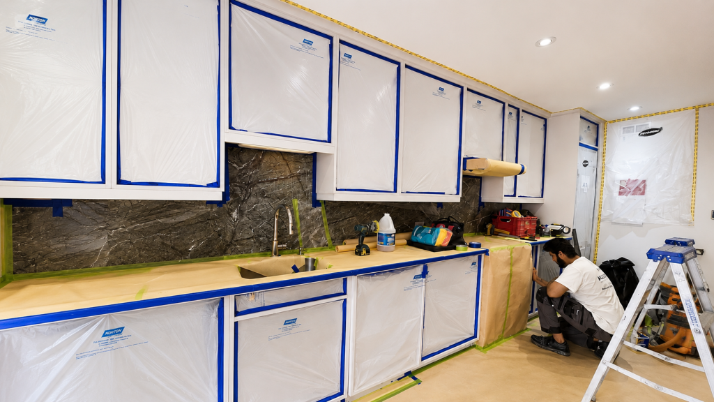 Worker crouching in a newly renovated kitchen, cabinets wrapped in protective plastic and blue painter's tape, with tools and a ladder nearby under construction lighting.